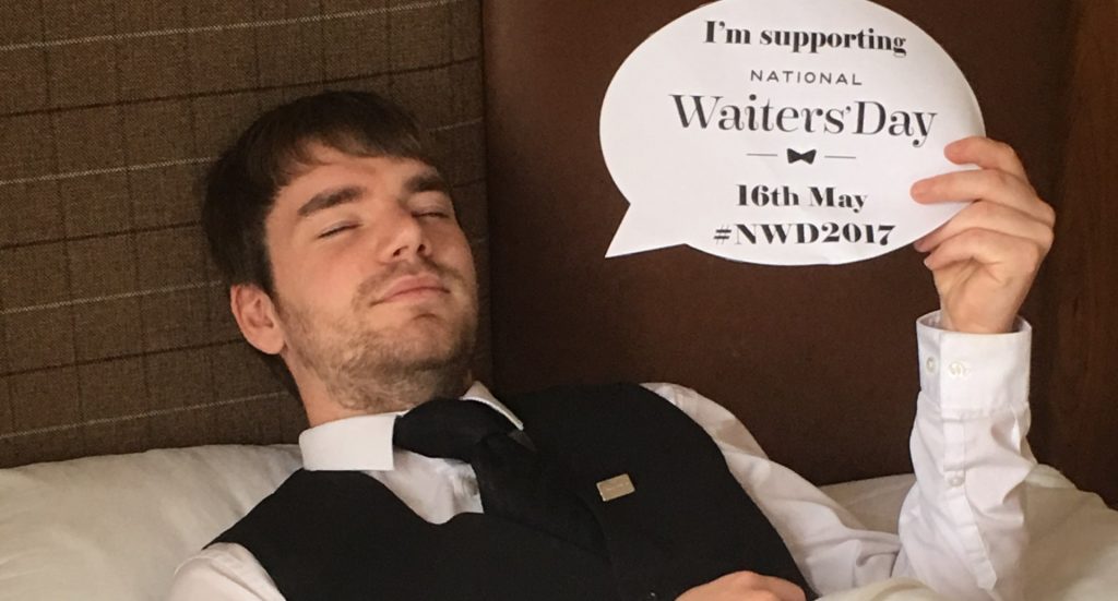 A young hotel waiter lies in a bed at Mackays Hotel with a sign saying "I support national waiters day"