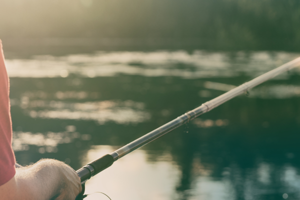 fishing rod close up with a man fishing into a river with the sunrise in front of him.