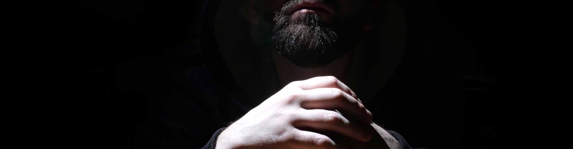 A bearded man shrouded in a hood holds his hands together.