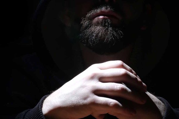 A bearded man shrouded in a hood holds his hands together.