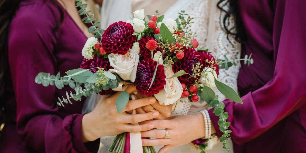 Bride and bridesmaids in elegant dress holding in hands bouquets of pastel burgundy flowers dahlia and greens eucalyptus with ribbon at nature. Young beautiful girls hold wedding bouquet outdoors.